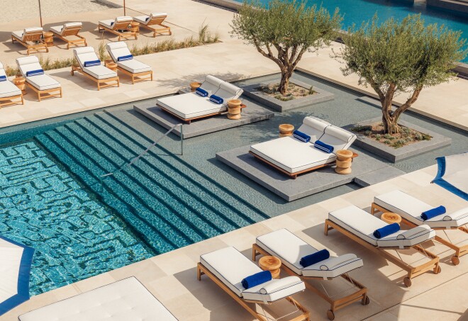 An overhead shot of a bright blue pool lined with white-cushioned wooden lounge chairs and olive trees 